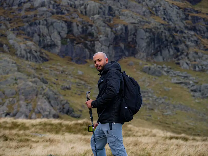 Man in a dark shell jacket hiking with a backpack and trekking pole across rocky mountain ground