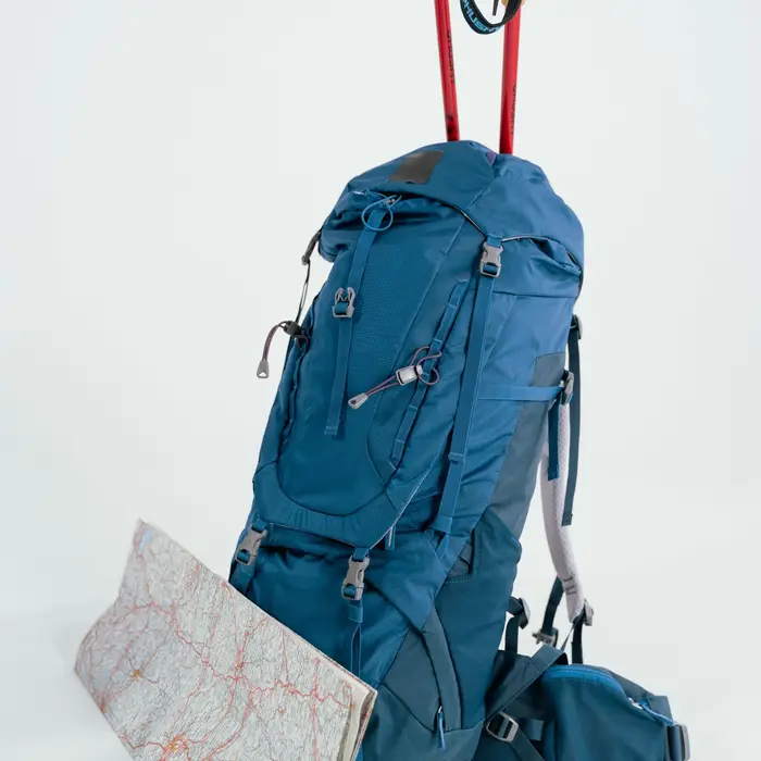 Blue hiking pack with trekking poles and a folded map on a light backdrop