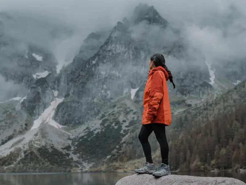 Woman in an orange shell jacket standing on a boulder by a misty mountain lake